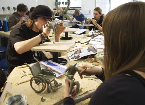 Students engaged in sculpture-making, focusing on creating figures from clay, with references and tools placed on shared workspace tables. One student is sculpting a character seated in a wheelchair, while another shapes a figure.