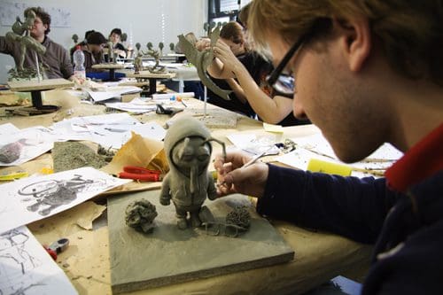 A student sculptor focuses on detailing a clay figure of a character wearing a hoodie at a workspace covered in sketches, tools, and clay materials, while classmates work on their own sculptures in the background.