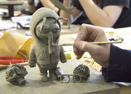 A sculptor working on a clay figure wearing a hoodie, featuring exaggerated facial features and antennae. Nearby are two small clay objects resembling broccoli and a pair of eyeglasses on a wooden work surface.