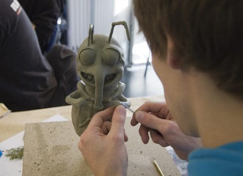 A person sculpting a cartoonish insect figure from clay, focusing intently on the details. The figure features large eyes and a whimsical expression, resting on a textured surface. In the background, another person is visible, engaged in a similar activity.