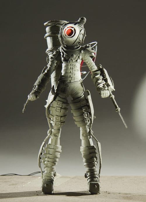 A stylized, futuristic character wearing a sleek, armored suit with a round helmet and a glowing red eye. The figure is holding two weapons and stands on a textured surface, illuminated by soft lighting to highlight the intricate details of its design.