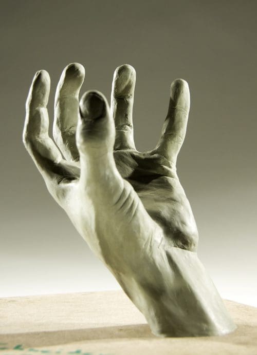 A sculpted human hand reaching upward, showcasing intricate details in the fingers and palm, with a smooth, gray surface. The background is softly blurred, emphasizing the hand's form.