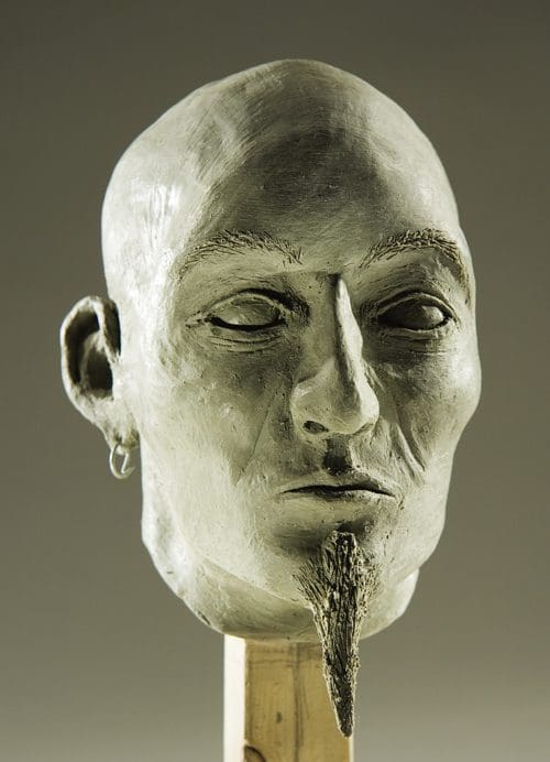 A sculpted head of a man with a smooth bald head and pronounced facial features, including a goatee and subtle ear decoration. The piece rests on a wooden stand against a neutral background, highlighting its artistic details.