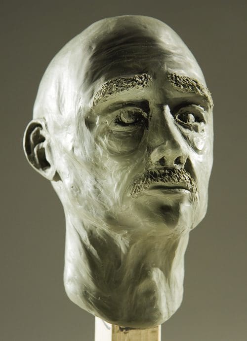 A detailed clay sculpture of a male head with prominent features, including a thin mustache and deep-set eyes. The head is slightly turned to one side, showcasing the textures and contours of the sculpted face.