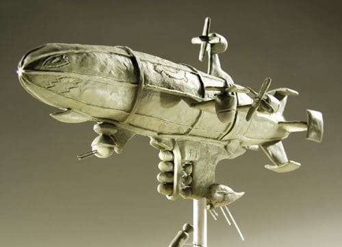 A detailed sculpture of a stylized airship with multiple propellers and features resembling a fish. The artwork has a metallic finish and is displayed on a stand against a soft, gradient background.