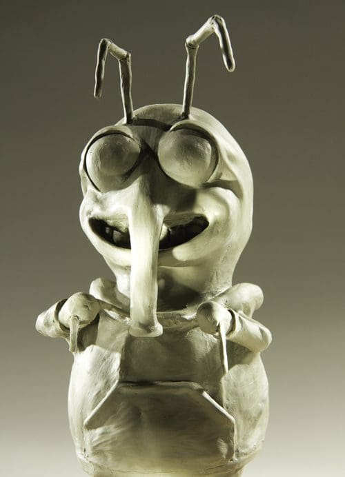 A whimsical clay sculpture of a character with large round glasses, an elongated nose, and antennae on top of its head. The figure has a playful smile and rounded body, showcasing a unique artistic style with a neutral background.