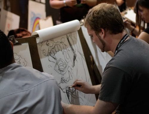 A man sketching on a large blank paper mounted on a stand, surrounded by others engaged in similar activities. The scene captures the creative atmosphere of an art-focused event, with various artwork in the background.