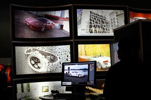 A person sitting at a computer surrounded by large screens displaying high-resolution images of luxury cars and artistic visuals, including a silver watch and abstract designs.
