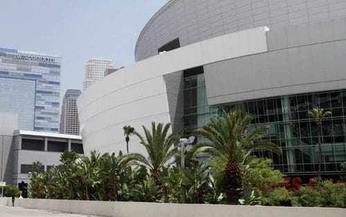 The exterior of a modern building featuring curved white architecture, complemented by lush palm trees and shrubs in the foreground. Skyscrapers and city buildings are visible in the background under a clear sky.