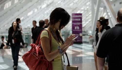 A woman in a yellow dress stands in a busy indoor space, focused on her smartphone. She carries a large red bag and has short, dark hair. In the background, people mingle and move about, with geometric patterns of light filtering through windows.