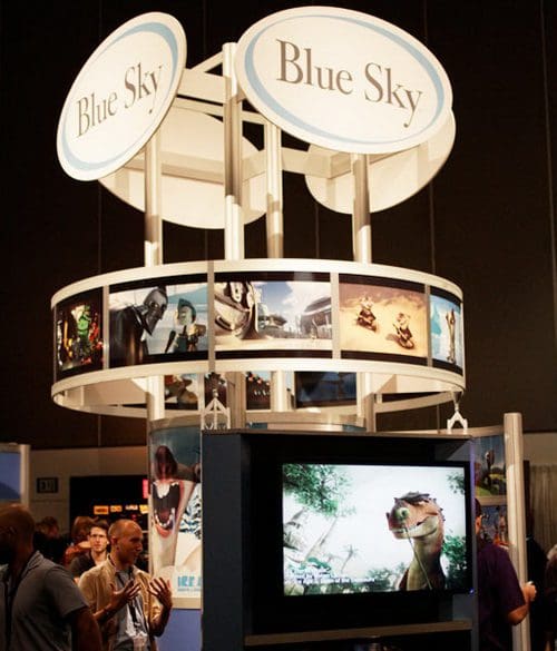 A trade show booth for Blue Sky Studios featuring a circular overhead sign and images from animated films displayed around the structure. Attendees are gathered nearby, with a large screen showcasing a character from one of the films.