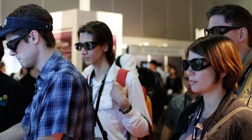 A group of four individuals wearing sunglasses at an event, engaged in different activities. The foreground features a man in a plaid shirt focusing on a touchscreen display, while two others observe. The atmosphere suggests a tech exhibition.