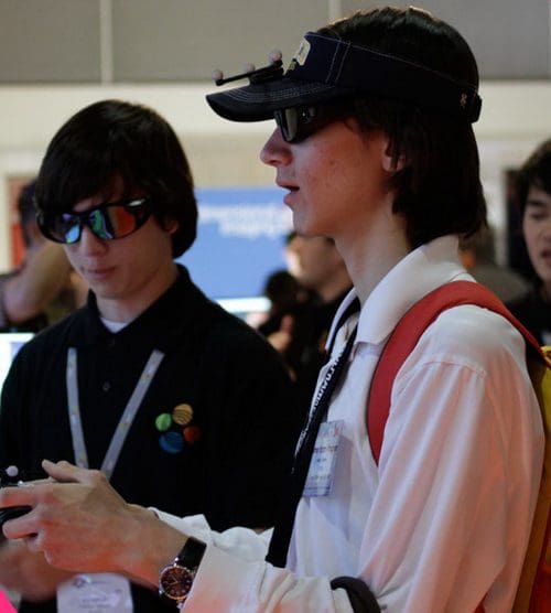 Two young men interacting at an event. One wears a black shirt with colorful buttons and sunglasses, while the other, dressed in a white shirt with a red backpack, appears focused on a handheld device. A blurred background suggests a busy exhibition atmosphere.