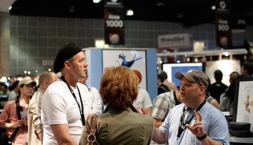A group of people engaged in conversation at a busy event. Two men, one in a beret and the other in a light blue shirt, are animatedly discussing with a woman in a green jacket, surrounded by a crowd and exhibition booths.