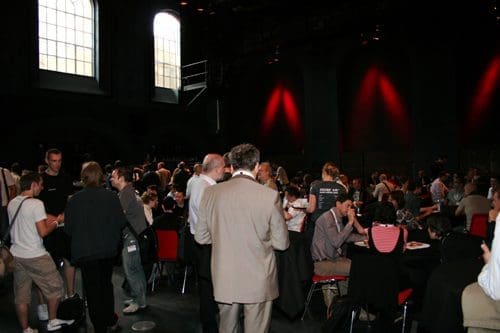 A large group of people mingling and engaging in conversation at a conference. The venue features high ceilings, large windows, and tables with attendees seated and networking.