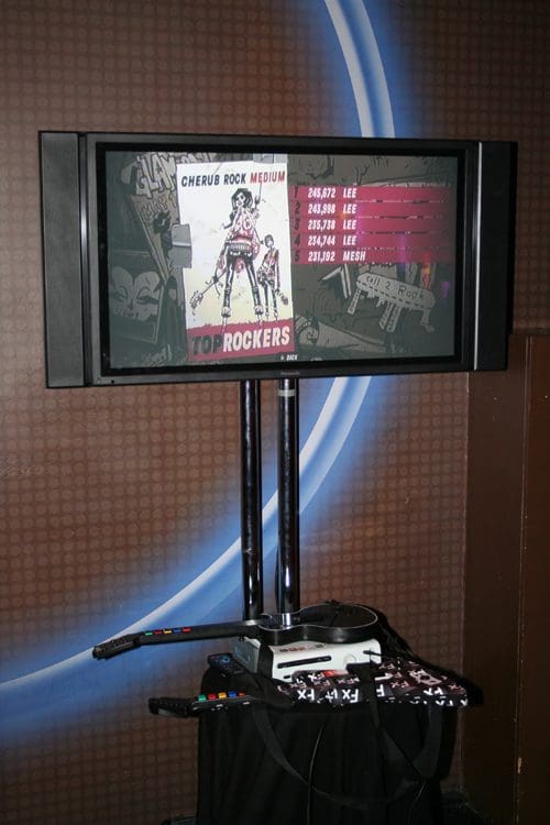 A large flat-screen monitor displaying a video game menu for a music game called 'Cherub Rock' with colorful graphics. Below the screen, a gaming console and a guitar controller are positioned on the floor.