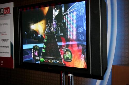 A large flat-screen television displays a video game interface with vibrant graphics. The screen shows colorful notes scrolling along a guitar neck, indicating gameplay in a music rhythm game.