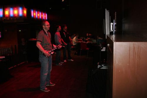 A man playing a video game with a guitar controller in a dimly lit room. He wears glasses and a striped shirt. In the background, others stand and sit, engaging with gaming stations and laptops.