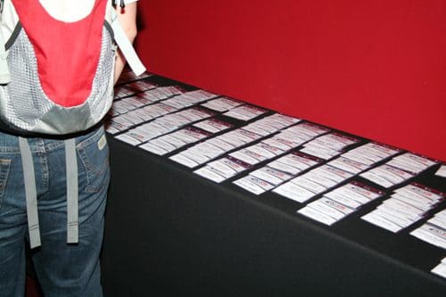 A person with a red and gray backpack stands next to a long table covered with numerous name tags arranged neatly. The background is a rich red color, creating a contrasting atmosphere.