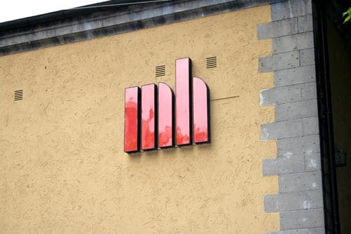 A modern logo displayed on a beige wall, featuring three stylized vertical bars with a gradient of red hues, suggesting a contemporary design aesthetic.