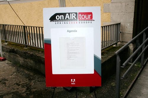 A large sign featuring a white background with red and black accents announcing the "on AIR tour Spring 2008 Europe," displaying an agenda in the center. The sign is placed outside against a textured wall.