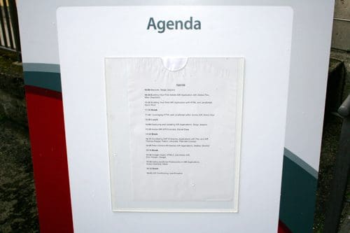 A weathered agenda displayed in a plastic sleeve, detailing the schedule of events. It is mounted on a signpost with a gray and red design, situated outdoors with blurred greenery in the background.