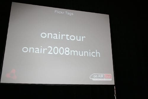 A projection screen displaying the text "Flickr Tags" with hashtags "onair tour" and "onair2008munich" in large font. The background is dark, enhancing the visibility of the text.
