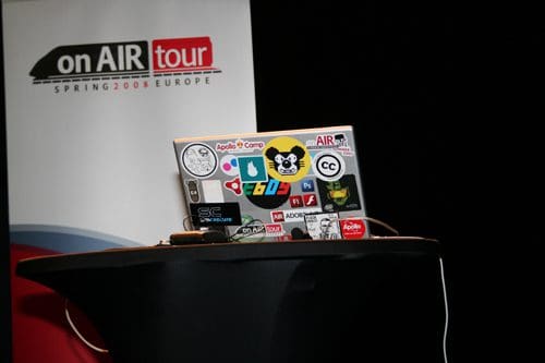 A laptop covered with various colorful stickers is placed on a black table, with a backdrop displaying "on AIR tour Spring 2008 Europe" in bold letters.