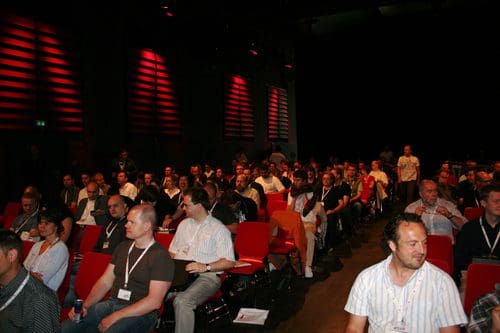 An audience seated in a dimly lit auditorium, facing a stage. Rows of people are engaged, with a mix of individuals in professional attire and name badges visible. The background features soft red lighting and dark walls.