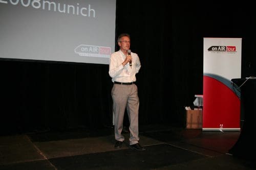 A man speaking at a microphone on stage, wearing a white shirt and gray trousers. Behind him, a projection screen displays text, and a banner on the side features a logo and branding.