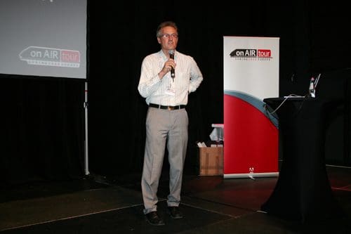A man with glasses stands on stage holding a microphone, delivering a speech. He is dressed in a light blue shirt and beige trousers. In the background, a banner for the 'on AIR tour' is visible, alongside a black podium.