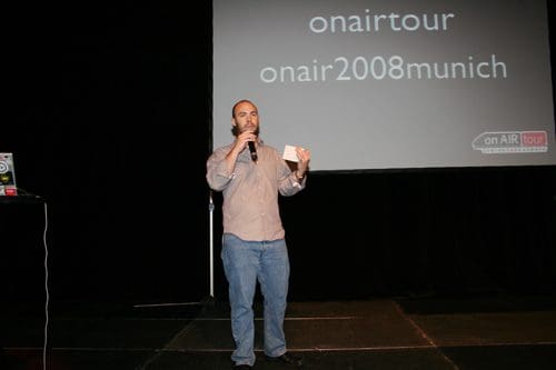 A man holding a microphone stands on a stage, speaking to an audience. He wears a light gray shirt and blue jeans, with a presentation backdrop displaying the text "onair tour" and "onair2008munich."