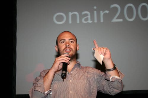 A man with a shaved head and beard speaking into a microphone at an event. He is wearing a gray button-up shirt and gesturing with one hand while holding a piece of paper in the other. A projection screen behind him displays the text 'onair200'.