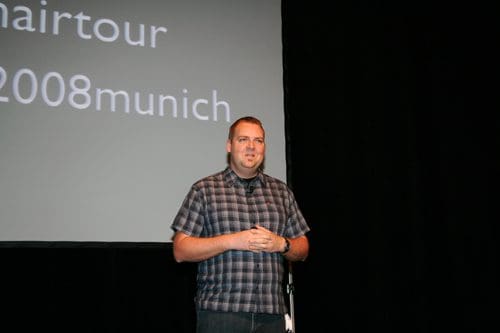 A man standing on stage in front of a presentation screen. He wears a checkered shirt and appears to be speaking to an audience. The background features text related to a 2008 event in Munich.