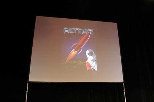 A presentation screen displaying a retro-style poster titled "ASTRO," featuring a large red rocket and an astronaut in a white suit with red accents. The backdrop depicts a celestial landscape with stars.