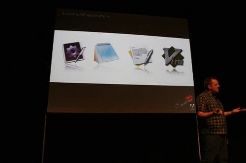 A presenter stands on stage in front of a large screen displaying various graphics software icons, including a graphic design program, a notepad, a pen, and a code editor, all arranged in a row.