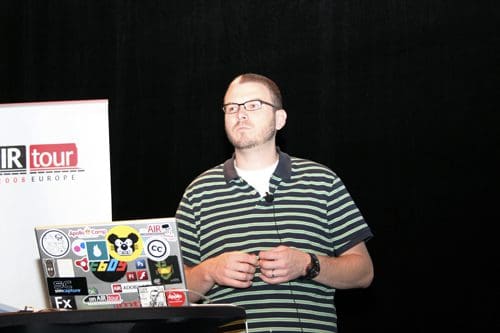 A speaker presenting at a conference, standing at a podium with a laptop covered in various colorful stickers. He is wearing a striped black and green polo shirt and glasses, with a thoughtful expression, against a dark background.