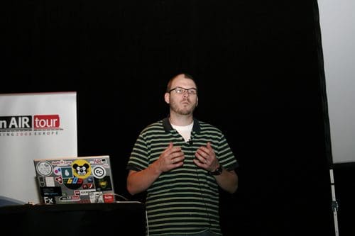 A speaker gesturing while presenting at an event, wearing a striped green and black polo shirt. A laptop covered in stickers sits on the table in front of him, with a banner in the background displaying the words 'AIR tour'.