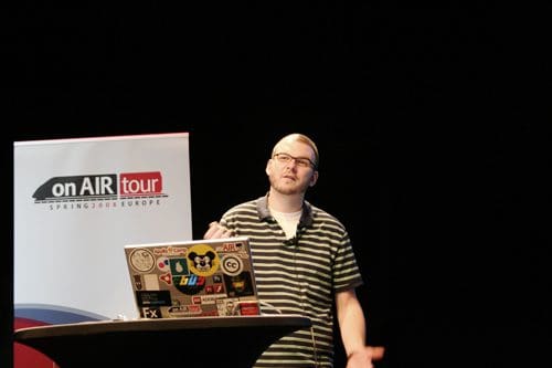 A speaker gesturing while presenting at a conference. He is wearing glasses and a striped black-and-white shirt, standing in front of a laptop covered with stickers on a podium. A banner that says 'on AIR tour' is visible in the background.