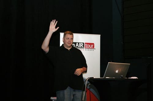 A man wearing a black shirt raises his hand during a presentation in front of a backdrop featuring the text 'AIR tour' and logos. A laptop is placed on a table nearby.