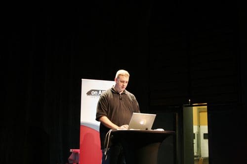 A speaker stands at a podium with a laptop during a presentation on stage, with a banner displayed in the background and a dimly lit theater setting.