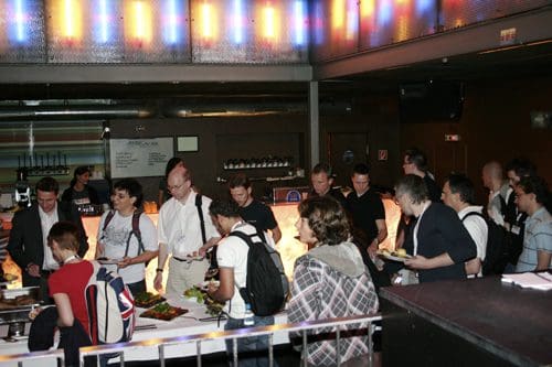 A diverse group of people, including men and women, standing in line at a buffet in a modern venue. They are engaged in conversation while selecting food from a colorful spread of dishes on a white counter.