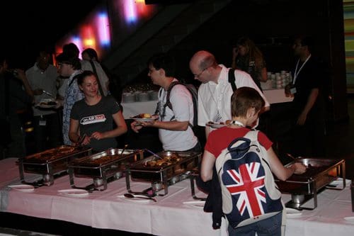 A buffet setup in a modern venue, with attendees serving themselves food from chafing dishes. One woman with a backpack featuring the Union Jack is smiling as she fills her plate, while others engage in conversation in the background.