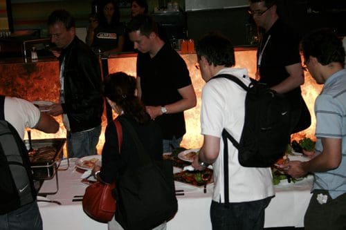 A group of people serving themselves at a buffet in a dimly lit venue. They are filling plates with food while standing near a table decorated with an orange glow. Some individuals carry backpacks and casual clothes.