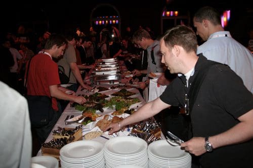 A group of people serving themselves at a buffet table filled with a variety of dishes and fresh vegetables. Several plates are stacked on the table in front of them as attendees choose food.