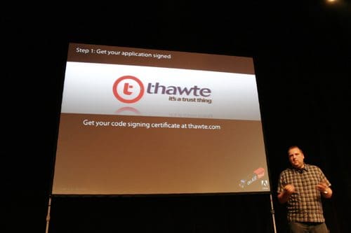 A speaker stands on stage in front of a large screen displaying instructions on obtaining a code signing certificate from Thawte. The slide emphasizes the importance of signing applications, featuring the Thawte logo.