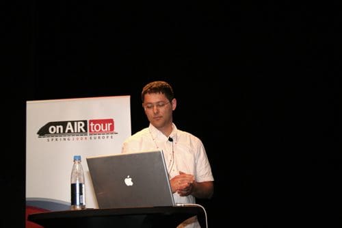 A speaker in a white shirt stands beside a laptop on a podium, presenting at an event. A banner with the words "on AIR tour" is visible in the background, indicating the theme of the presentation.