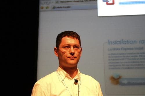 A man wearing glasses and a white shirt stands in front of a large screen displaying software installation instructions. He looks focused with a neutral expression, while the bright screen content is partially visible.
