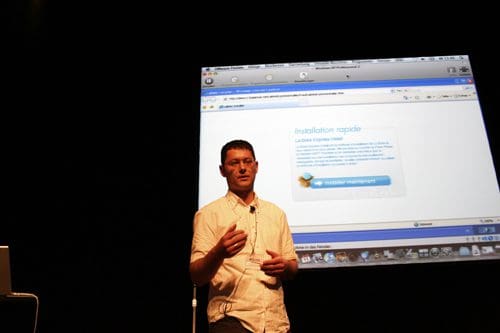 A speaker presenting on stage in front of a large screen displaying a web installation process. The speaker gestures with one hand while holding a microphone, wearing a light-colored shirt, with a focused expression.