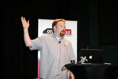 A man gesturing with his hand while speaking at a podium during a presentation. He has a beard and wears a plaid shirt. A laptop is open on the table beside him, with a sign in the background featuring the text 'on tour'.